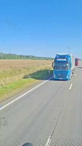 The video shows a view from inside a vehicle traveling on a road. Initially, a large grey trailer truck is visible in the foreground, with another red truck and trailer passing by in the distance. The road is asphalt with white lane markings. The surrounding environment consists of fields and vegetation under a clear blue sky. As the video progresses, the grey truck is no longer in view, and the perspective shifts to show more of the road ahead, with the red truck now further in the distance. The camera continues to move forward, and a single white car is seen in the distance on the road. Roadside markers with red and white stripes are visible along the edge of the road.