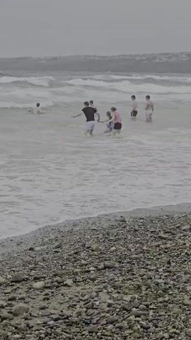 The media file shows a group of people at a beach in Tramore, Ireland. The scene is a moderately wavy ocean with a pebble beach in the foreground. There are several individuals in the water, appearing to be enjoying themselves despite the overcast and potentially cool weather. Two people in the foreground are wading into the water, one wearing a black shirt and shorts, the other a pink top and dark shorts. Further out, more people are visible in the waves, some of them appearing to be children. The overall impression is of a casual beach outing, possibly on a day with less than ideal weather, but where people are still determined to enjoy the water. The time of day appears to be daylight, given the ambient light, but the sky is overcast, suggesting a cloudy or potentially rainy day. The location is clearly identified as Tramore, Ireland. There is no visible text in the media file.