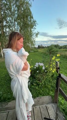 A woman wearing a white robe is standing on a wooden deck and holding a baby wrapped in white fabric. She is looking to her left. In front of her, to her right, is a phone on a tripod. Beyond the deck is a grassy area with some flowers. Further in the background, there is a body of water with a small structure on it, surrounded by trees and a field. The sky is visible with clouds, and the sun appears to be setting, casting a warm glow. The woman says "Come out, damn it, come on. Listen, it's closing, not your camera."