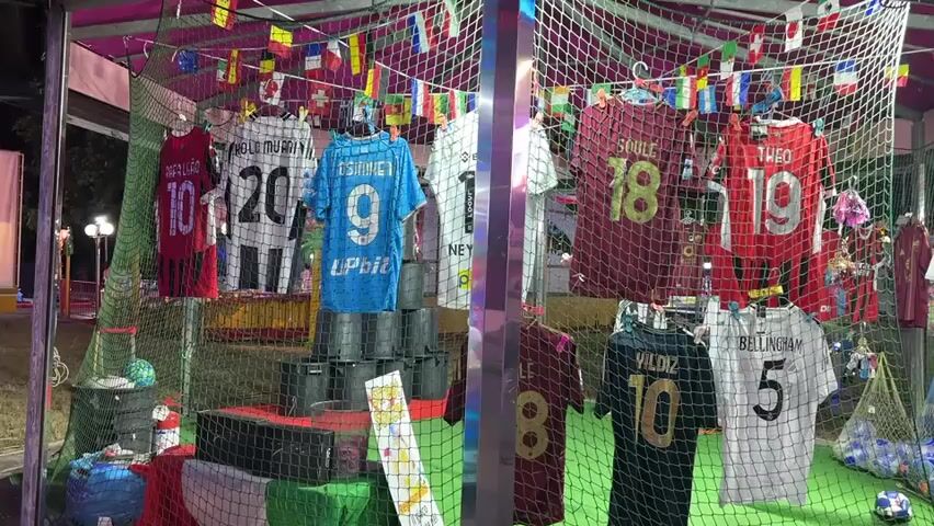 The video pans across a stall displaying soccer jerseys for sale at an outdoor event, possibly a fair or carnival, at night. The stall is adorned with small national flags strung across the top and is enclosed by netting. Several jerseys with player names and numbers are hanging on display. On the left, red, black and white, and blue jerseys are visible. On the right, maroon and red jerseys are displayed. Below these, more jerseys are hung, including black and white ones. In the background, people are seen walking around, and illuminated attractions of the event are visible. There are also soccer balls in netting containers. A few individuals, mostly young males, are gathered around the stall, appearing to be customers or spectators. The lighting suggests it is evening or night, with artificial lights illuminating the scene.