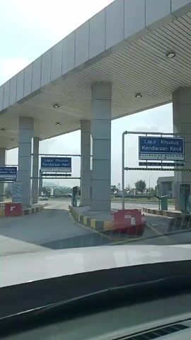 The video shows a view from inside a vehicle approaching a toll plaza. The camera is looking forward through the windshield. The toll plaza has multiple lanes, with overhead signs indicating "Lajur Khusus Kendaraan Kecil" which translates to "Special Lane for Small Vehicles". There are also signs indicating a maximum height of 2.1 meters. The lanes are marked with yellow and black striped barriers. To the right of the lane closest to the camera, there is a green toll payment machine and a fire extinguisher mounted on a pillar. The sky is bright and overcast.