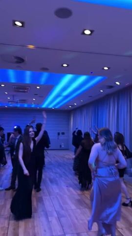 The video shows a group of young people dancing at a party. The room is lit with blue lights on the ceiling and recessed lighting. Most of the attendees are dressed in formal attire. The floor appears to be made of wood. People are moving and dancing with energy.