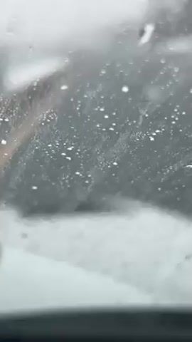 The video shows a view from inside a car during heavy snowfall. The windshield is covered with droplets and slush, obscuring the view of the road ahead. A yellow line is visible on the road, indicating a lane marker. The wipers are not activated.