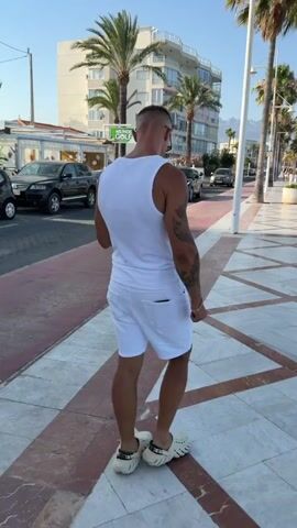 A man in a white tank top and white shorts walks on a sidewalk. He has short, dark hair and tattoos on his left arm. He is wearing white clogs. To his left, there is a street with cars parked and driving. In the background, there is a building with palm trees in front of it. To his right, there is a row of palm trees and a street lamp. The sky is clear and blue. The sun is shining.