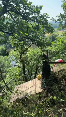 A young woman wearing a pink helmet and pink and grey harness is on a wooden platform in a forest. She is holding onto a zipline cable. She is looking at the camera and smiling. She is also wearing a light green fanny pack. Behind her, another person in a red helmet is partially visible on another platform. The background shows a river and rolling hills. The video is taken from a low angle.