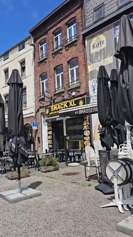 This video shows a view of an outdoor seating area for restaurants and bars on a sunny day. Tables and chairs are set up on a cobblestone or paved surface, with many black umbrellas closed. Some of the establishments have signage visible, including "DEEP HOUSE BAR" and "SNACK XL BRASSERIE," and a menu board is present. The buildings lining the street appear to be multi-story with a mix of architectural styles. There are a few people visible walking in the distance. The sky is blue with some clouds.