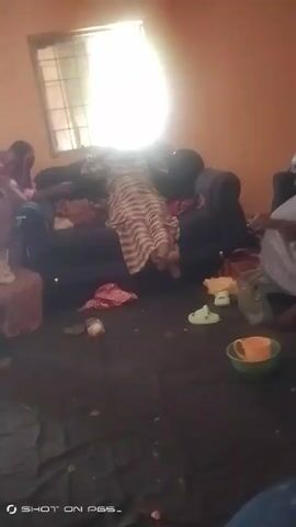 The video shows several people sitting in a room. There are three women on the sofa. One of them is lying down. Another woman is sitting in a chair beside the sofa. The floor is covered in dark material. Several items are scattered across the floor, including a bowl, shoes, and fabric. There is a window in the background. The room has orange walls. The camera moves to show a woman in a yellow dress. The scene is blurry and the lighting is poor.