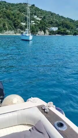 The video captures a serene daytime scene on clear blue waters in a coastal area. The perspective is from a small boat, with its white cushions and an inflatable fender visible in the foreground, suggesting the boat is either anchored or moving slowly.

Initially, an anchored sailboat with a tall mast is visible in the middle distance, surrounded by the calm, turquoise sea. Behind it, a lush green hillside rises, dotted with white buildings under a bright blue sky.

The camera then pans to the right, revealing a wider expanse of the sparkling blue water. A small motorboat with several people aboard can be seen navigating across the water further in the distance. The coastline continues, featuring more green hills that gradually transition into distant mountains, partially veiled by light clouds. The sky remains clear and bright, indicating good weather. The water has gentle ripples, reflecting the sunlight. The overall setting is a picturesque marine landscape.