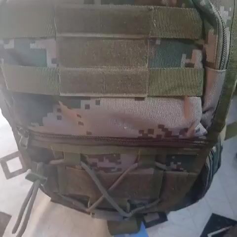 The video shows a military-style tactical backpack with a digital camouflage pattern. It has several MOLLE webbing straps on the front, a zippered compartment at the bottom, and a crisscrossed bungee cord. The backpack is likely sitting on a table or other flat surface, and there's a hint of a blue object in the background. The video seems to be taken indoors.