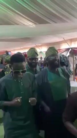 The video captures a scene at what appears to be a social event, possibly a wedding or party, under a white tent. Several people are present, mostly dressed in shades of green. In the foreground, a young man wearing sunglasses and a green shirt stands with his hands in front of him, looking around. Beside him is a bearded man in a matching green outfit and traditional cap. The camera moves slightly, revealing more of the crowd, including women in patterned dresses and headwraps, and other men in similar green attire. Some people are moving around, suggesting a lively atmosphere. Floral decorations and tables are visible in the background, adding to the festive ambience.