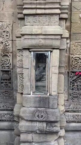 The video shows a close-up view of an ancient stone sculpture enclosed behind glass. The sculpture appears to be of a deity or mythical figure, with intricate carvings and details. The surrounding structure is also made of stone, with decorative elements and weathered surfaces, suggesting it is part of a historical temple or monument. The lighting is somewhat dim, and the glass reflects the surroundings, including trees and the sky, indicating the sculpture is likely outdoors.