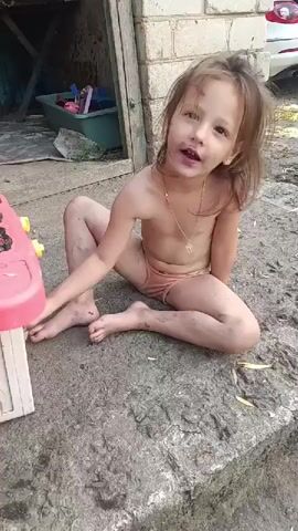 A young girl is sitting on a concrete surface in an outdoor setting. She appears to be very young, with long, light brown hair that is messy and appears to have dirt in it. Her face is also smudged with dirt, but she is smiling and looking directly at the camera. She is wearing light-colored underwear and a simple gold chain around her neck. Her legs and arms are also covered in dirt.

To the left of the girl, a portion of a pink and white toy is visible. In the background, there is a brick wall, some wooden structures, and what appears to be a green tub with items inside. A white car is partially visible in the far background. The ground around the girl is dusty and appears to be made of dirt and concrete. The lighting suggests it is daytime, with sunlight casting shadows. The overall scene conveys a sense of playfulness and perhaps a bit of mischievousness, as the girl seems to have been playing in the dirt. The location context is Saksahan, Ukraine. There is no visible text in the media file.
