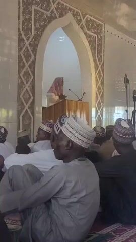 A low-angle shot depicts a group of men in a mosque, sitting on the floor and facing a speaker behind a wooden podium. The speaker, draped in a light-colored garment, appears to be addressing the gathering. The mosque's interior features white walls with decorative arches and geometric patterns around the podium area. The men are wearing a variety of traditional caps and clothing, with some in white and others in darker shades. A camera on a tripod stands to the side, possibly recording the event. The ceiling has a series of circular light fixtures. The room is well-lit, possibly with natural light streaming in from windows that are visible in the background.