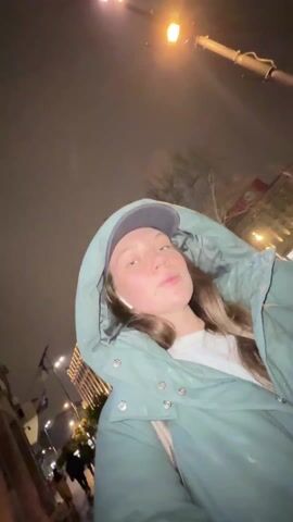 A young woman stands outside at night, under a streetlight. She wears a green hooded jacket with a cap underneath. She has earbuds in and appears to be looking around. The scene includes city buildings and some light precipitation, possibly rain or snow.