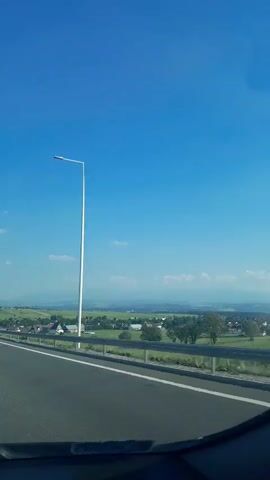 The media file shows a view from a moving vehicle on a highway in Lasek, Poland. The scene is set during the daytime, under a clear blue sky with a few scattered white clouds. The weather appears to be pleasant and sunny.

The main subjects are the highway, a roadside guardrail, and a distant landscape. The highway has clear white lane markings and a shoulder. A tall white lamppost stands on the side of the road, a common sight on highways. Beyond the guardrail, the landscape features a rural scene with scattered houses, green fields, and trees. In the far distance, there are rolling hills or mountains, softened by a slight haze.

There is a green road sign visible at the 00:01 mark of the video, indicating "Łyse 3" and "Rybno 6". This suggests the vehicle is traveling on a route leading towards these locations.

The overall mood is one of travel and observation, with the camera capturing the passing scenery. The focus is on the journey and the expansive view, rather than specific people or detailed objects within the vehicle. The clear weather and bright daylight contribute to a sense of openness and freedom associated with road trips.