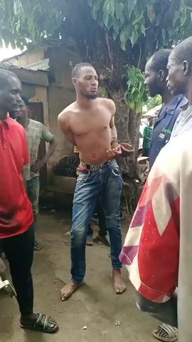 In an outdoor setting in Kaduna South, Nigeria, a group of men is gathered around a shirtless man in blue jeans, who stands barefoot with his hands behind his back, suggesting he is restrained. He has a distressed or pleading expression on his face, indicating he is the focus of the group's attention.

Several other men surround him: one in a red polo shirt stands on the left, while another in a blue work-type shirt with "Barber" visible on the chest stands on the right. An older man in a patterned shirt is next to the man in the "Barber" shirt. Other individuals are visible in the background, some wearing striped shirts. The ground is dusty, and simple structures with corrugated iron roofs and leafy trees are visible in the background, suggesting a village or informal urban area. The lighting indicates it is daytime, possibly overcast or shaded. The shirtless man's belt has "MEN" written on it. The overall atmosphere appears tense, suggesting a confrontation or questioning is taking place.