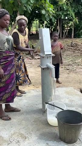 The video shows three people standing around a manual water pump. A woman on the left is wearing a striped shirt and a colorful skirt. A woman in the center is wearing a yellow shirt with a pattern and a patterned skirt, and she is operating the pump. A young boy is standing to the right of the pump, looking towards the side. Two buckets are placed near the pump to collect water. The scene appears to be outdoors, with trees and foliage in the background. The ground is muddy.