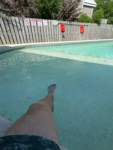 A first-person perspective video shows a person's leg submerged in shallow water in a swimming pool. The pool is outdoors, with a wooden fence and buildings visible in the background. The water is clear and the sun is shining. The video pans slightly to the left and then back to the right.
