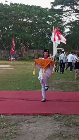 A young person in a white uniform and a black hat walks on a red carpet outdoors. They are holding a folded orange fabric with yellow tassels. The person is marching with one leg raised in a parade-like manner. In the background, there are trees, a white wall, people watching, and flags, including a white and red flag. The ground is covered in grass and dirt.