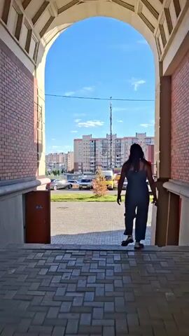 A woman with long, dark hair walks down stairs through an archway. She is wearing a dark blue dress. The scene is outdoors, with a clear blue sky and white clouds. In the background, there are several apartment buildings and a yellow construction crane. A red sign with a yellow "M" logo and the Russian word "Магнит" (Magnit) is visible on the right side of the frame. Cars are parked on the street in front of the buildings. The sun is shining, casting shadows on the ground, suggesting it is daytime. The overall impression is a typical urban scene on a sunny day.
