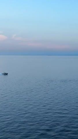 A small boat travels across a vast, calm body of water under a clear, pale blue sky streaked with soft pink clouds. The boat is in the lower left quadrant of the frame and is moving from left to right, leaving a white wake behind it. The sun is low in the sky, creating a serene and tranquil atmosphere. In the distance, the faint outlines of land or other vessels are visible on the horizon. The water is a deep, tranquil blue, reflecting the colors of the sky. The overall scene is one of peaceful solitude and vastness.