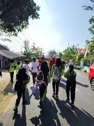 A group of people, appearing to be mostly children and young adults, are walking down a paved road. The sun is shining, casting long shadows. The people are dressed in various casual clothes. Some of the children are wearing bright green vests with numbers on them. One person in the center is wearing a white t-shirt with the number 92 on it. The group is walking away from the camera, heading towards buildings and vegetation visible in the distance. A flag, which appears to be the Indonesian flag, is visible in the background. The road is lined with houses and trees. The overall impression is of a group participating in an outdoor activity or event.