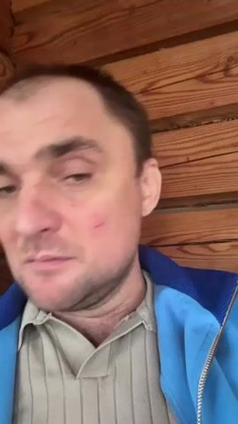 A close-up video shows a man speaking to the camera. He has fair skin, brown hair, and a beard. There are scratches on his left cheek. He is wearing a blue jacket over a gray patterned shirt. He speaks, then smiles, and then speaks again. The background is a wall made of horizontal wooden planks.