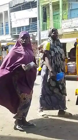 Here's a description of the video:

The video shows two women walking on a street in what appears to be an urban area. One woman is wearing a purple hijab and a colorful patterned skirt, holding a metal plate. The other woman is wearing a gray and white patterned hijab, carrying a child on her back and a blue plastic bottle in her hand.

They walk towards a yellow auto rickshaw, where a man in a black outfit gets in as a passenger. Another man in a red shirt is visible inside the rickshaw. The background features buildings, street signs, and other people.