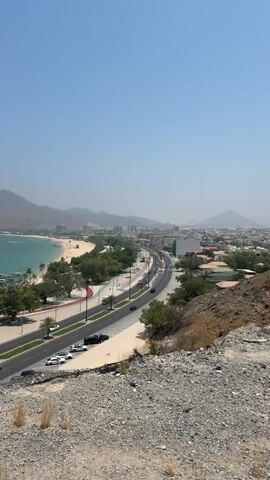 An elevated, panoramic view of a coastal city on a clear, sunny day. The camera slowly pans from right to left, revealing a multi-lane highway running parallel to a sandy beach and the blue-green ocean. Palm trees and greenery line the road and beach. In the mid-ground, a city with various buildings extends along the coast, backed by mountains on the left. As the camera continues to pan, a distant cargo port with several container ships and large cranes becomes visible on the horizon over the water. The immediate foreground shows dry, rocky terrain and a dark metal railing on the lower left. Multiple vehicles are observed moving and parked on the highway.