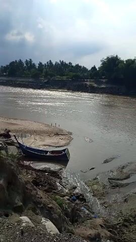 The video pans right across a muddy river towards a bridge. A small boat is docked on the left bank of the river. The water is brown. The bridge is constructed with a metal lattice and is decorated with red and white flags. There are palm trees on the banks of the river. The sky is mostly clear.