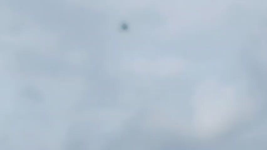 A small airplane is seen in the sky against a cloudy backdrop. The plane is a dark silhouette against the lighter sky and appears to be flying in a straight line. The image is a still frame from a video, with timestamps indicating it was captured around the beginning of the video.