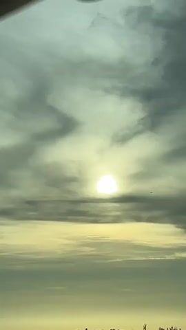 The video shows a cloudy sky with the sun partially visible. Below the sky is a horizon with a line of trees and some silhouetted structures. The sun is bright and creates a yellowish glow. There is a small airplane visible in the distance. The bottom of the video shows the top of a vehicle, indicating the footage is likely taken from a moving car. The overall scene appears to be a landscape view, possibly during sunrise or sunset.
