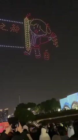 The video shows a drone light show against a night sky. At the start, the drones are arranged to form an image of a character holding a scroll, alongside a line of text in Chinese characters. The drones then reposition to display the year "2026". The light show is viewed by a crowd gathered on a tiered, illuminated staircase. The cityscape is faintly visible in the background. The crowd is seen holding up phones, presumably to record the spectacle.