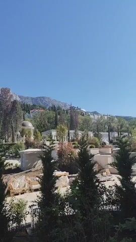 The video shows a park landscape with workers and construction materials. In the background, a mountain range stretches across the horizon, and a tall communications tower stands to the left. The foreground is filled with trees, including several evergreen trees, and some materials for a building project, including wooden pallets and wrapped items. Several people are visible in the park, one of whom appears to be working with tools. The sky is clear and blue, suggesting a bright, sunny day.