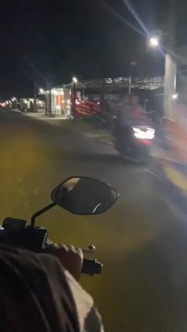 A first-person perspective video shows a person riding a motorcycle at night on a paved road. The motorcycle's headlight illuminates the road ahead with a yellow glow. On the right side of the road, there are several buildings, including a red and yellow bus and what appear to be small shops or stalls. Streetlights are visible along the road. The rider's hand is visible on the handlebar, and a rearview mirror reflects the rider's face and the surroundings. The overall environment is dark, suggesting a rural or suburban setting at night.