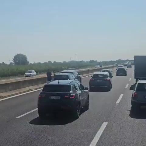 A drone shot slowly pans over a highway with moderate traffic. Several cars are visible, most of which are SUVs and sedans. One man is seen walking between two cars on the highway. The environment appears to be rural, with fields and trees visible beyond the highway. The sky is clear and the weather seems to be sunny.