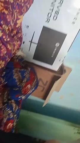 A video shows a partially opened box for a curved monitor. The box is mostly white with black text and an image of the monitor. The brand name "IMMER" is visible on the box. To the right of the monitor box, a brown cardboard box is also visible. A person's hand is seen briefly at the bottom of the screen. The background appears to be a wall with some light blue paint and beige splotches. There is also some fabric with a floral pattern visible in the foreground. The video is shaky and blurry.