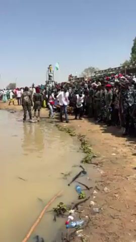 The video shows a crowd of people gathered along a muddy riverbank on a sunny day. On the left, some people wade in the shallow water. A man in a white shirt walks near the edge of the water. A group of people wearing white vests with red cross symbols carries a stretcher with a person lying on it through the crowd. There are uniformed security personnel standing on the right side of the crowd. In the background, a structure with a Nigerian flag on top can be seen. The ground is covered with mud, plastic bottles, and some green plants. The video seems to capture a crowded event with potential medical attention being provided.
