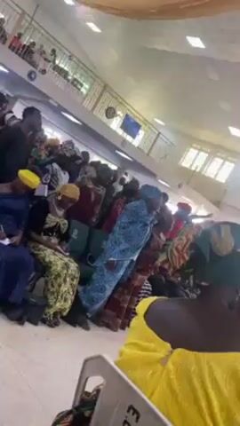 The video shows a gathering of people inside a well-lit building, likely a church or auditorium. The majority of the individuals are women, many of whom are wearing colorful traditional African attire, including headwraps. Some are seated and appear to be writing in notebooks. There are also a few young men present, some of whom are standing and interacting with the seated women. The camera angle is from a low perspective, looking up at the crowd, with a partial view of the ceiling, which features multiple light fixtures. The atmosphere seems to be one of reverence or attentiveness.