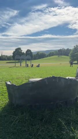 A video pans across a grassy park on a sunny day with a blue sky and scattered clouds. In the foreground, a dark-colored, ornate sculpture resembling a sofa or chaise lounge is visible, partly obscured by grass. As the camera moves, it reveals more sculptures of chairs, also dark-colored and ornate. In the middle ground, there is a paved path and a set of three arched structures. Further in the background, more chairs are placed in front of the arches, and there are trees and rolling hills. A person in dark clothing is visible standing near the trees on the right side of the frame. The overall environment appears to be an outdoor art installation or sculpture park.