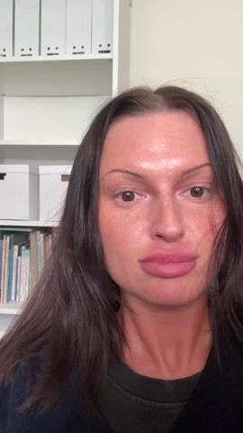 The video starts with a close-up shot of a woman with long, dark hair, fair skin, and prominent lips. She is wearing a dark-colored sweater. Behind her is a white bookshelf filled with books and white storage boxes. She smiles slightly and asks if the viewers want to see what she does in a day. The camera abruptly switches to a dark red screen, then shifts rapidly to show a computer screen, possibly a monitor, partially obscured by white blinds. The movement is quick and slightly shaky.