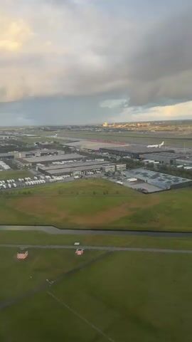 An aerial view captures an airport complex under a dramatic sky with clouds. In the foreground, a grassy field features navigation markers and a small red and white building. Further into the frame, several large industrial buildings with flat roofs are visible, some with numerous trucks parked in designated areas. A narrow waterway snakes through the landscape. In the background, airport runways stretch out, with a distant airplane visible on one. The overall scene suggests a functional, operational airport environment bathed in the soft light of either sunrise or sunset.
