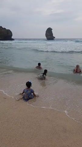 The video shows a beach scene with people in the water. A young girl in a blue floral dress initially sits on the sand facing the ocean. She then stands up and moves closer to the water. Further in the water are three other individuals, including a woman wearing a hijab. The waves are small and the sky is overcast. In the background, there are rocky formations both in the water and along the shoreline. The beach sand is light tan.