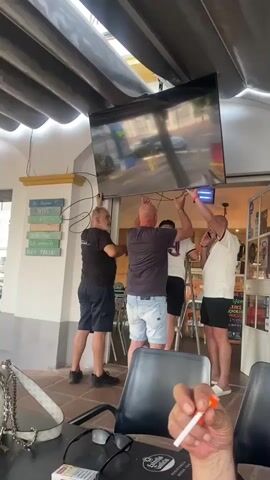 Four men are working together to install a large flat-screen television. Two men are on the left, one wearing a black t-shirt and shorts, the other in a dark grey t-shirt and denim shorts. Both are holding the television from below, with their arms extended upwards. One of them is standing on the ground, while the other is on a stepladder. Two other men are on the right, both also holding the television from below. One man is wearing a white t-shirt and black shorts, and the other is wearing a white t-shirt and dark shorts. The television is angled upwards and appears to be connected by several cables. The men are in an outdoor covered area, possibly a patio or balcony, with a tiled floor and a white awning above. There are signs on a pillar to the left, and a doorway to the right leads into a building. In the foreground, a person's hand is visible holding a cigarette.