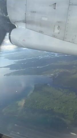 The video shows an aerial view from a small plane, with part of the propeller visible in the frame. Below, there's a landscape with a network of waterways, likely rivers or deltas, interspersed with islands and land covered in dense green vegetation. The water appears to be a mix of blue and cloudy hues.