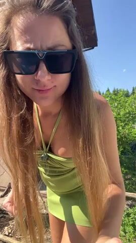 A pregnant woman in a green dress and sunglasses stands outside a log cabin, in front of trees and a blue sky. She holds her stomach and looks toward the camera. Music plays in the background.