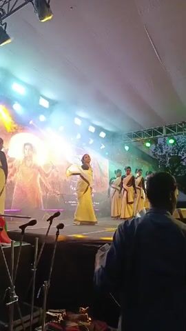 The video captures a stage performance with several people. In the foreground, a person in a blue jacket faces away from the camera, looking towards the stage. The stage itself is illuminated by bright lights and smoke effects.

On the stage, a central figure, dressed in a white sari with a dark blouse, is performing a dance. They are holding a white fabric, extending it with their arms. To the right and slightly behind this dancer, a group of about seven individuals, also in similar white saris and dark blouses, are lined up, facing the audience.

In the background, a large screen displays an image, possibly a backdrop for the performance. The image on the screen features a central figure in warm tones, surrounded by abstract designs and text in a non-English script. Stage lights, including spotlights and green lights, are visible above the stage. Some microphones on stands are positioned on the stage floor in the foreground. The overall environment appears to be an outdoor or semi-outdoor event, given the presence of trees visible in the upper right corner.