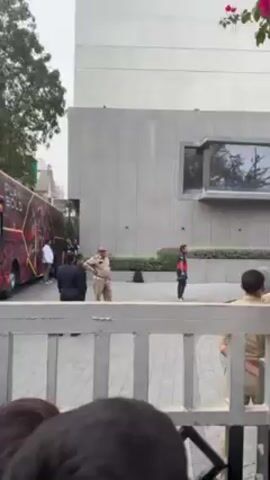 The video shows a group of people standing outside a modern building with a "Play Bold" bus parked nearby. Several police officers are present, some standing guard and others in conversation. A person with a camera stands off to the side. The view is partially obstructed by a white gate. The scene seems to be a security detail possibly waiting for people to enter or exit the building. The weather appears to be overcast.