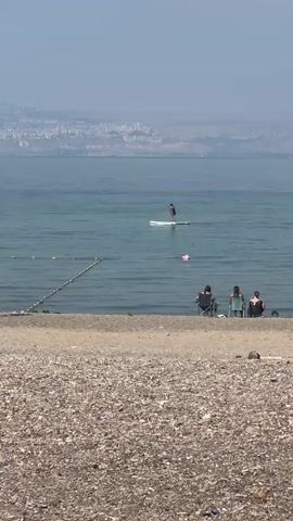 The video captures a serene beach scene. In the foreground, a pebbly beach leads to the calm waters of a large body of water, possibly a lake or sea. A rope barrier with yellow buoys separates the shallow swimming area from the deeper water. In the middle ground, three people are seated in beach chairs on the shore, facing the water. Further out, a lone individual stands on a paddleboard, actively paddling. In the background, a hazy cityscape stretches across the distant shoreline. The overall atmosphere is tranquil and sunny.