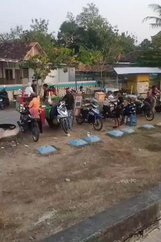 A group of people are gathered outdoors. Several motorcycles are parked nearby. There are a few small stalls set up, one appearing to sell food or drinks from a cart. People are interacting around the stalls, and some are seated on the ground. In the background, there are buildings with tiled roofs and trees. The overall environment suggests a casual gathering or market in a rural or semi-rural setting. The lighting indicates it may be late afternoon or early morning.